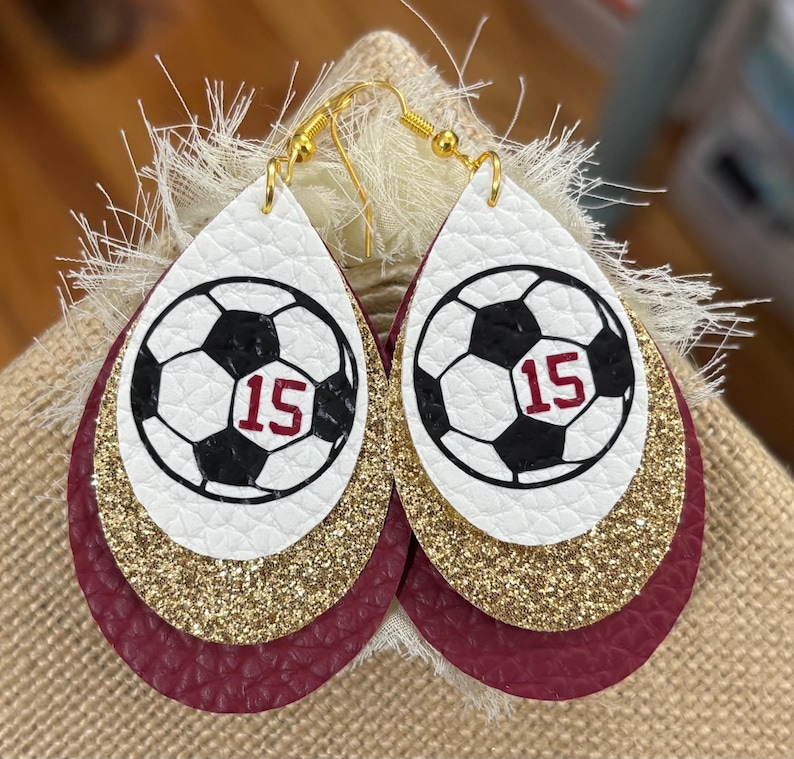May include: Teardrop-shaped earrings with a soccer ball design. The earrings feature a white top layer with a black and white soccer ball graphic and the number 15 in red. Gold glitter and burgundy leather layers complete the design.
