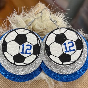 May include: A pair of round earrings featuring a soccer ball design with the number 12 in blue. The earrings have a white and black soccer ball design, layered over silver glitter and blue glitter. The earrings hang from silver hooks.