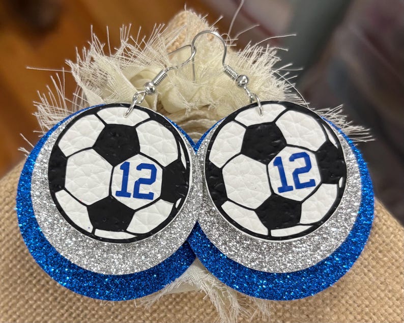 May include: A pair of round earrings featuring a soccer ball design with the number 12 in blue. The earrings have a white and black soccer ball design, layered over silver glitter and blue glitter. The earrings hang from silver hooks.