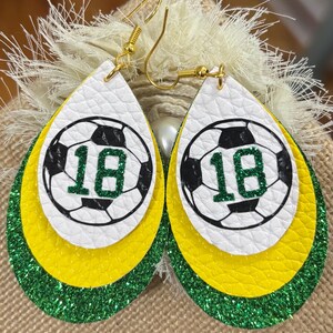 May include: Teardrop-shaped earrings with a soccer ball design. The earrings feature a white top layer with a black and white soccer ball graphic and the number 18 in green. The middle layer is yellow, and the bottom layer is green glitter.