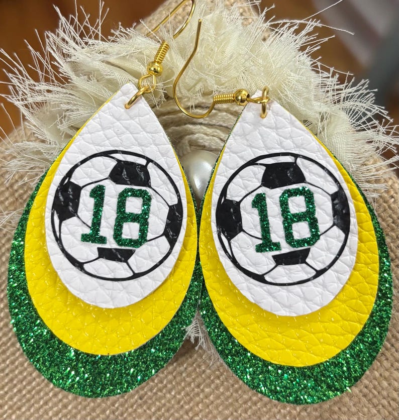 May include: Teardrop-shaped earrings with a soccer ball design. The earrings feature a white top layer with a black and white soccer ball graphic and the number 18 in green. The middle layer is yellow, and the bottom layer is green glitter.