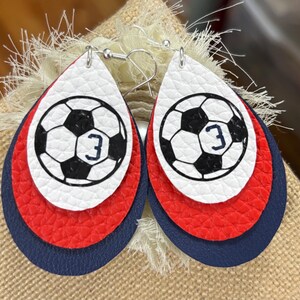 May include: A pair of layered teardrop earrings. The top layer is white with a black and white soccer ball design and the number 3. The middle layer is red, and the bottom layer is navy blue. The earrings have silver-colored hooks.