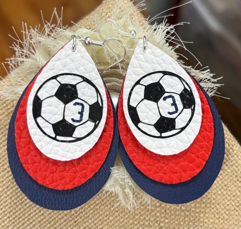 May include: A pair of layered teardrop earrings. The top layer is white with a black and white soccer ball design and the number 3. The middle layer is red, and the bottom layer is navy blue. The earrings have silver-colored hooks.