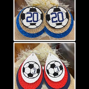 May include: Two pairs of soccer-themed earrings. One pair features a soccer ball design with the number 20, set against blue and gold glitter layers. The other pair has a soccer ball with the number 3, layered with red and navy blue.