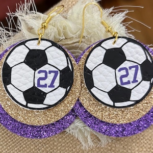 May include: A pair of soccer ball earrings. Each earring features a white and black soccer ball design with the number 27 in purple. The earrings have a gold glitter layer and a purple glitter layer. The earrings hang from gold-colored hooks.