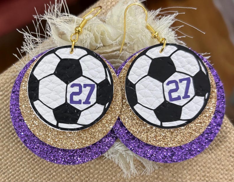 May include: A pair of soccer ball earrings. Each earring features a white and black soccer ball design with the number 27 in purple. The earrings have a gold glitter layer and a purple glitter layer. The earrings hang from gold-colored hooks.