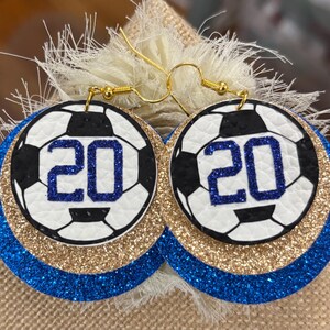May include: Soccer ball earrings with the number 20 in blue glitter. The earrings have a white and black soccer ball design, gold glitter, and blue glitter layers. The earrings hang from gold-colored hooks.