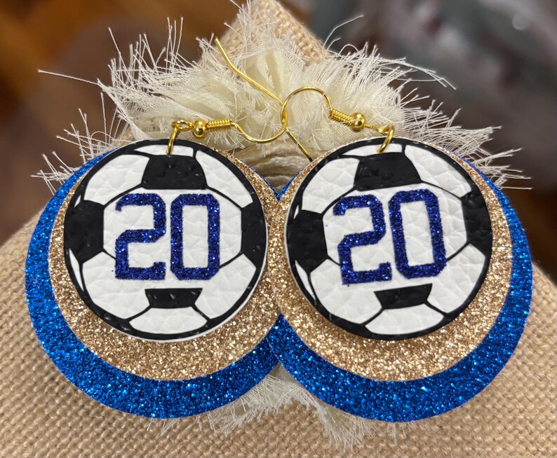 May include: Soccer ball earrings with the number 20 in blue glitter. The earrings have a white and black soccer ball design, gold glitter, and blue glitter layers. The earrings hang from gold-colored hooks.