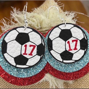 May include: A pair of soccer ball earrings. Each earring features a black and white soccer ball design with the number 17 in red. The earrings are layered on red and blue glitter circles, with silver hooks.
