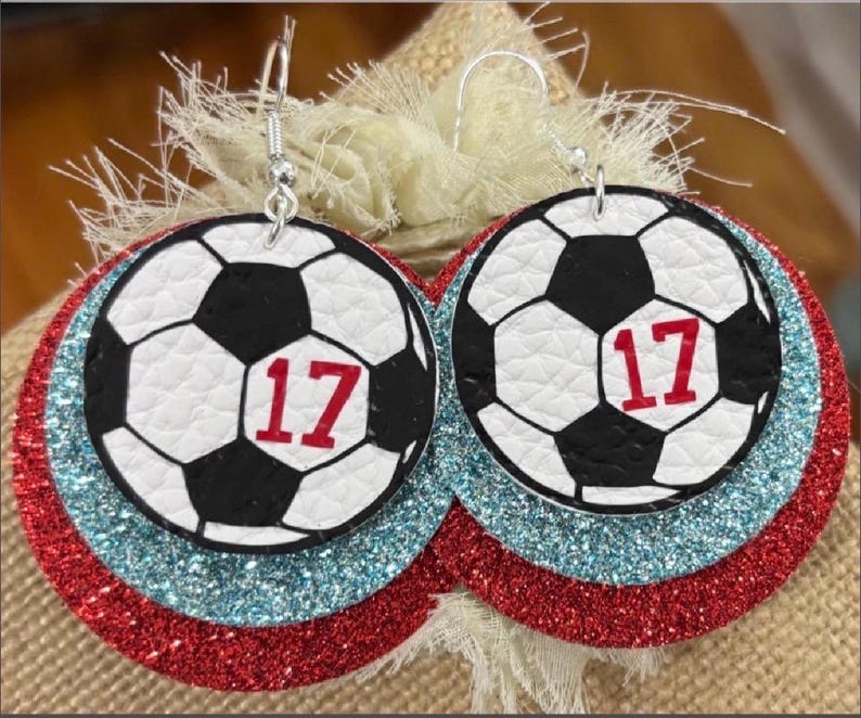 May include: A pair of soccer ball earrings. Each earring features a black and white soccer ball design with the number 17 in red. The earrings are layered on red and blue glitter circles, with silver hooks.