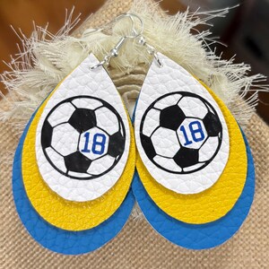 May include: A pair of layered teardrop earrings. The top layer is white with a soccer ball graphic and the number 18 in blue. The middle layer is yellow, and the bottom layer is blue. The earrings have silver hooks.
