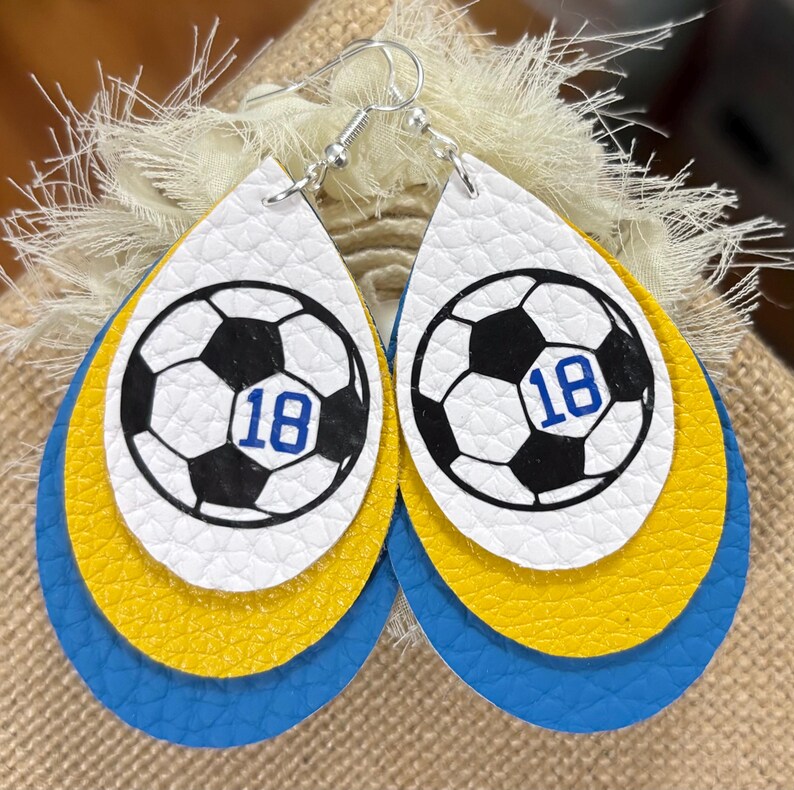 May include: A pair of layered teardrop earrings. The top layer is white with a soccer ball graphic and the number 18 in blue. The middle layer is yellow, and the bottom layer is blue. The earrings have silver hooks.
