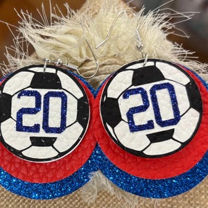 May include: A pair of round earrings with a soccer ball design. The earrings feature a white background with a black soccer ball pattern and the number 20 in blue glitter. The earrings have red and blue glitter layers.