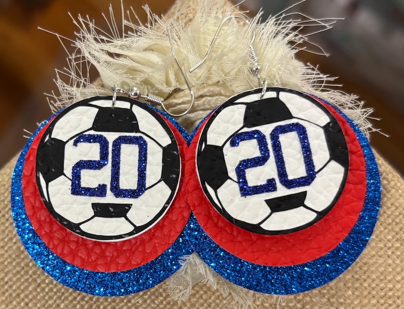 May include: A pair of round earrings with a soccer ball design. The earrings feature a white background with a black soccer ball pattern and the number 20 in blue glitter. The earrings have red and blue glitter layers.