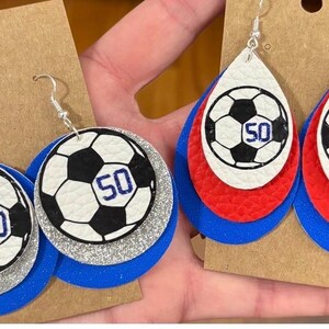 May include: Soccer ball earrings with a black and white design and the number 50 in blue. The earrings are layered with blue, silver, and white or red, white, and blue. The earrings hang from silver hooks.