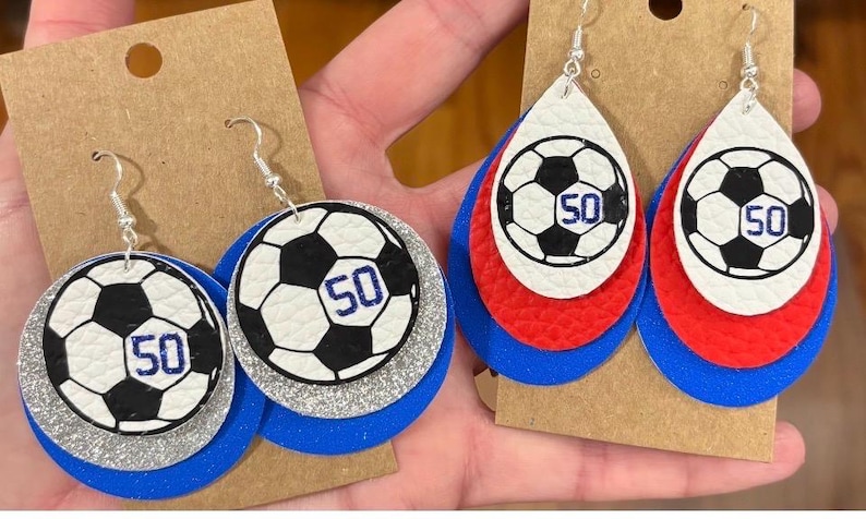 May include: Soccer ball earrings with a black and white design and the number 50 in blue. The earrings are layered with blue, silver, and white or red, white, and blue. The earrings hang from silver hooks.