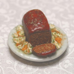 Könnte beinhalten: Miniatur-Hackbraten mit roter Glasur, serviert auf einem weißen Teller mit geschnittenem Gemüse. Der Hackbraten ist dunkelbraun, und das Gemüse ist weiß und orange. Der Teller hat einen hellblauen Rand.