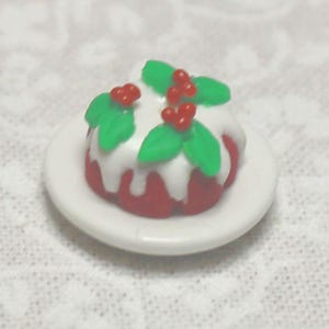 May include: A miniature red cake with white frosting, green leaves, and red berries, presented on a small white plate. The dessert is a festive decoration, possibly for a dollhouse or holiday display.