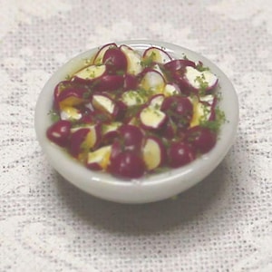 May include: A miniature white bowl filled with a salad. The salad contains sliced red radishes, yellow pieces, and green herbs. The bowl is set on a white, textured surface.