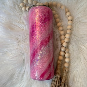 May include: A pink and white glitter tumbler with a swirl design. The tumbler is on a white fluffy surface with a wooden bead garland and a brown tassel.