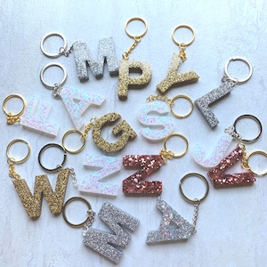 May include: A collection of 12 sparkly letter keychains in various colors, including silver, gold, pink, and white. Each letter is made of resin and has a glitter finish. The keychains are attached to silver key rings.