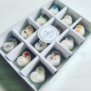 May include: A white box containing 12 heart-shaped wax melts. The box is divided into 12 compartments, each holding a different coloured wax melt. The wax melts are decorated with glitter. The box has a label that reads "The Cottage Wax Melts".