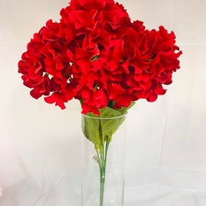 May include: A bouquet of vibrant red artificial flowers in a clear glass cylinder vase. The flowers have multiple layers of petals, and the stems are visible through the glass. The vase is approximately 12 inches tall.