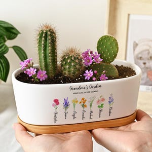 May include: An oval white ceramic planter with a bamboo base containing two cacti and small pink flowers. The planter features the text Grandma's Garden, Make Life More Grand, and a series of floral illustrations with names printed underneath each flower.