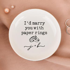 Może przedstawiać: Biała ceramiczna miseczka na pierścionki z napisem "I'd marry you with paper rings" i dwiema ilustracjami pierścionków. Widoczne są również dwa srebrne pierścionki i naszyjnik z pereł. Miseczka stoi na różowej powierzchni.