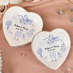 Peut inclure: Deux petits plats en céramique en forme de cœur, blanc cassé, avec une bordure dorée. Chaque plat présente une illustration bleue sur le thème du mariage et le texte "Kalyah & Marry 06.18.2025".