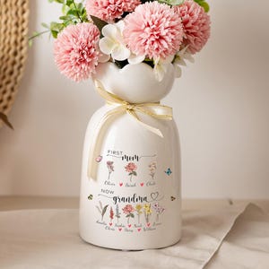 Op de afbeelding: Een witte keramische vaas met een geschulpte rand en een crèmekleurig lint om de hals. De vaas is voorzien van bloemillustraties en de tekst "FIRST mom" en "NOW grandma" met diverse namen gedrukt onder de bloemafbeeldingen.