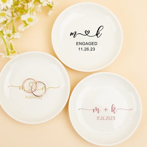 May include: Three white ceramic ring dishes with gold and pink lettering. The first dish has the text "m & k" with a heart between the letters and the date "11.23.2023". The second dish has the text "m & k" with a heart between the letters and the text "ENGAGED" above the date "11.26.23". The third dish has the text "m + k" with the date "11.26.2023" below.