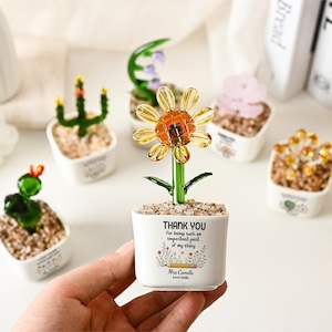 May include: A glass sunflower figurine in a white ceramic pot filled with small stones. The pot features the text: THANK YOU for being such an important part of my story, a pencil illustration, and the name Mrs. Camila Love Stella.