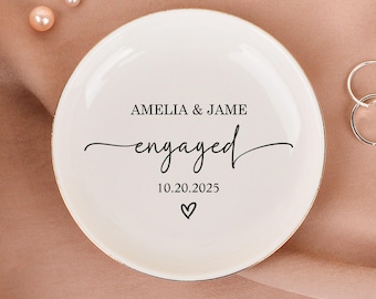 Könnte beinhalten: Eine weiße Keramik-Ringschale mit den Worten "AMELIA & JAME engaged 10.20.2025" in schwarzer Schrift. Die Schale ist von Perlen, Ringen und Blumen auf rosa Hintergrund umgeben.