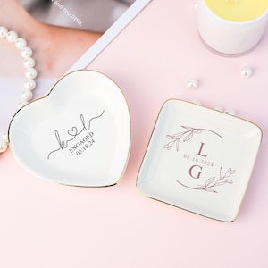 Może przedstawiać: Dwa białe ceramiczne talerzyki na pierścionki z pozłacanym wykończeniem. Jeden talerzyk ma kształt serca i napis "k&l ENGAGED 05.18.24". Drugi talerzyk jest kwadratowy i ma kwiatowy wzór z inicjałami "L" i "G" oraz datą "08.16.2024".