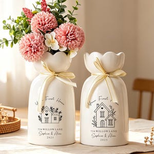 May include: Two white ceramic vases with scalloped rims, each tied with a cream ribbon. The vases feature a black line drawing of a house, the text "Your First Home," "724 WILLOW LANE," "Sophia & Alex," and "2025." One vase holds pink flowers.