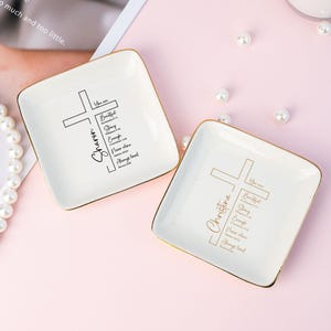 May include: Two square, white ceramic trinket dishes with gold rims. Each dish features a cross design and inspirational text. One dish says "Sharon" and the other says "Christina". Pearls and a pink background complete the scene.