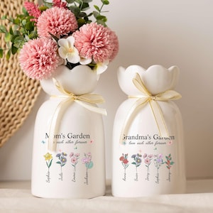 Puede incluir: Dos jarrones de cerámica blanca con arreglos florales y cintas doradas. Uno de los jarrones dice "Mom's Garden" y el otro "Grandma's Garden". Cada jarrón tiene un diseño floral y nombres. Los jarrones están sobre una superficie blanca.