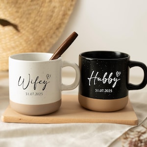 May include: Two ceramic mugs, one white and one black, with speckled patterns. The white mug says "Wifey" and the black mug says "Hubby" in script, with a date below. Both mugs have a tan base and are on a wooden tray.