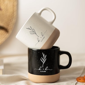 Puede incluir: Dos tazas de cerámica, una blanca y otra negra, apiladas una encima de la otra. La taza blanca tiene un acabado moteado y una base beige, mientras que la taza negra tiene un acabado moteado y una base beige. Cada taza presenta un diseño botánico y el texto "k + h 10.23.2025".