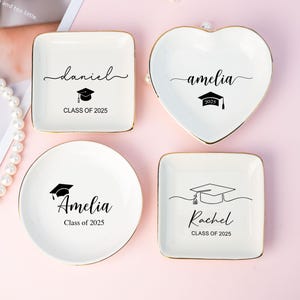 Puede incluir: Cuatro platos de cerámica blanca para joyas con ribete dorado. Cada plato tiene un diseño diferente con un gorro de graduación y el nombre "Daniel", "Amelia", "Amelia" y "Rachel" con el año "2025".