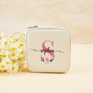 Pu&ograve; includere: Una scatola per gioielli bianca con chiusura a zip. La scatola ha un design floreale rosa con la lettera "S" e il nome "sophia" scritto in corsivo nero.