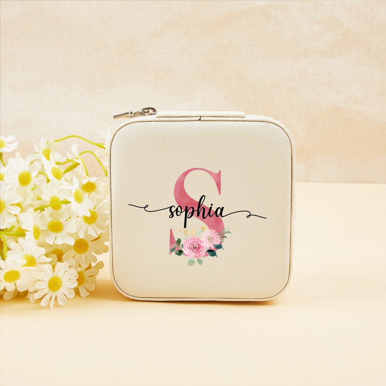 Pu&ograve; includere: Una scatola per gioielli bianca con chiusura a zip. La scatola ha un design floreale rosa con la lettera "S" e il nome "sophia" scritto in corsivo nero.