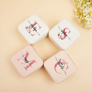 Pu&ograve; includere: Quattro scatole per gioielli quadrate con disegni floreali e iniziali personalizzate. Le scatole sono bianche e rosa con un motivo floreale e le iniziali sono in un font corsivo.