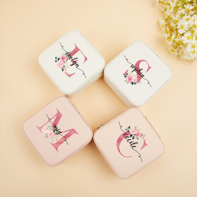 Pu&ograve; includere: Quattro scatole per gioielli quadrate con disegni floreali e iniziali personalizzate. Le scatole sono bianche e rosa con un motivo floreale e le iniziali sono in un font corsivo.