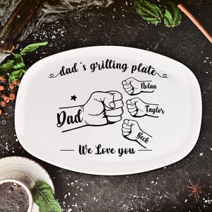 Puede incluir: Plato ovalado blanco para asar a la parrilla con texto negro que dice "dad's grilling plate" y "Dad". El plato presenta ilustraciones de puños y los nombres Nolan, Taylor y Beck. También se incluye el texto "We Love you".