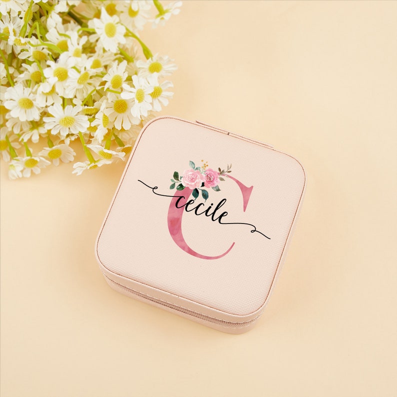 Pu&ograve; includere: Una scatola per gioielli rosa con chiusura a zip. La scatola presenta un design floreale con una lettera "C" rosa ad acquerello e la parola "Cecile" scritta in corsivo nero.