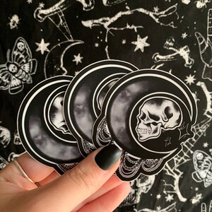 May include: A stack of black and white stickers featuring a skull inside a crescent moon design. The stickers are held against a black background with a repeating pattern of skulls, moths, and stars.