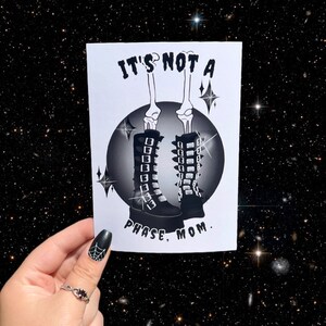 Puede incluir: Una tarjeta blanca con texto negro y una ilustración en blanco y negro de un esqueleto con botas negras con hebillas. El texto dice "No es una fase, mamá."