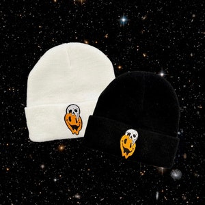 Könnte beinhalten: Zwei Strickmützen mit einem Totenkopf- und Smiley-Motiv. Eine Mütze ist weiß und die andere ist schwarz.